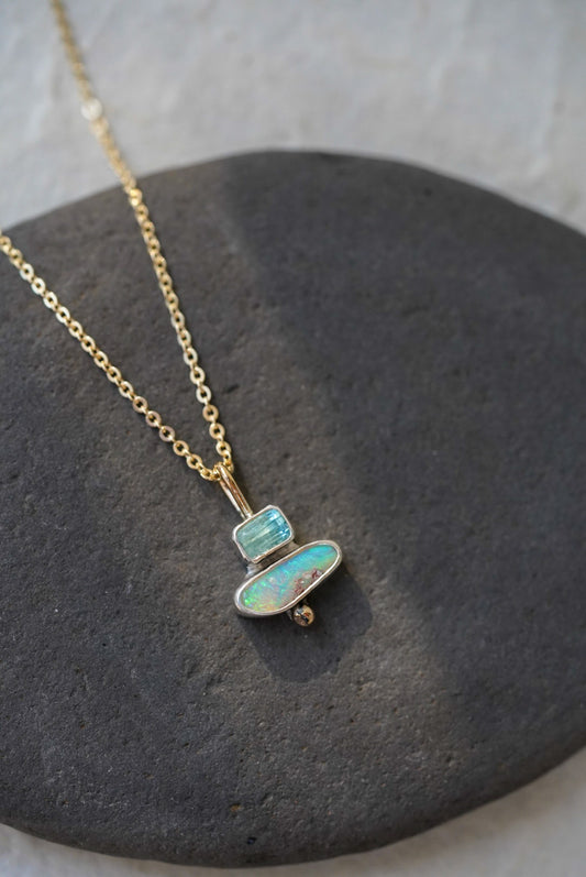 A delicate gold chain necklace with a pendant made of stacked natural stones, one tourmaline and one opal, displayed on a dark gray stone slab against a textured off-white background.