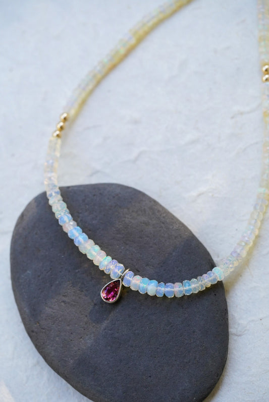 A opal strand beaded necklace accented with small gold beads and a dark pink teardrop tourmaline, displayed on a dark stone against a textured off-white background.