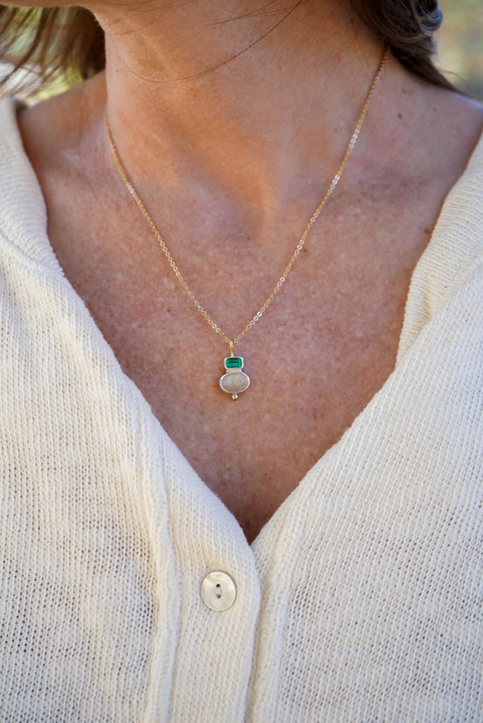 A close-up of a person wearing a delicate gold chain necklace with a small silver pendant featuring a green tourmaline on top and an opal on the bottom, styled with a cream-colored knit cardigan.