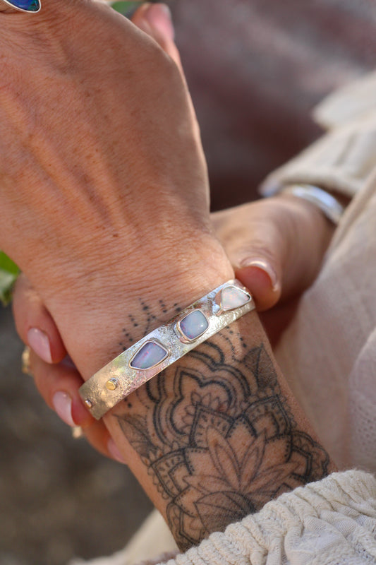 Close-up of a hand wearing a silver bracelet with three Australian Opal stones, held by another hand with tattoos.