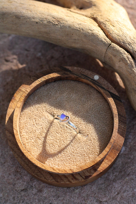 Wooden ring box with a heart shaped opal ring inside, placed on sand with driftwood.
