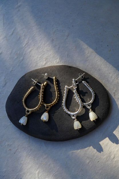 Two pairs of dangling hoop earrings displayed on a dark stone slab against a light textured background. One pair is bronze with hand carved flower bead drops, and the other is silver with hand carved flower bead drops. 