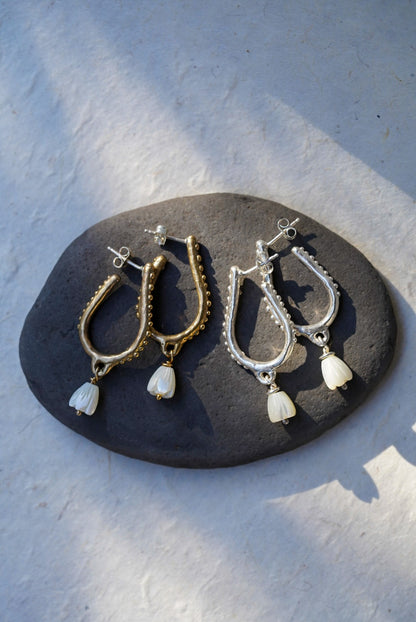 A pair of bronze and silver hoop earrings with hand carved flower pearl charms, displayed on a dark stone plate against a textured light gray background.