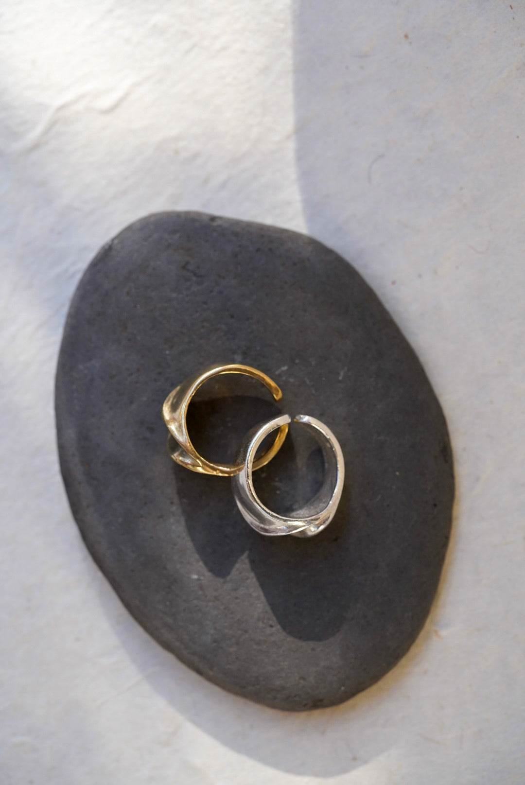 Two textured open rings, one bronze and one silver, displayed on a dark gray stone against a light textured background.