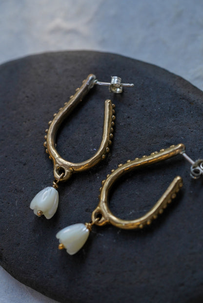 A pair of bronze hoop earrings with hand carved flower beads, displayed on a dark stone against a light gray textured background.