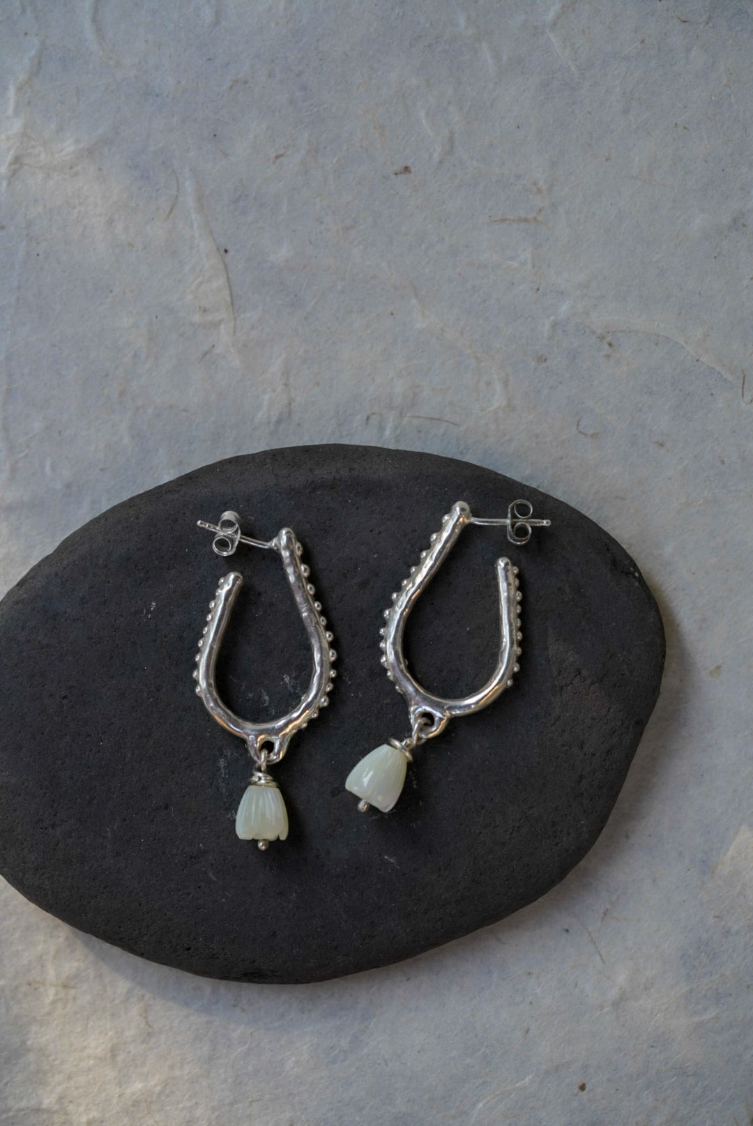 A pair of silver hoop earrings with hand carved flower beads, displayed on a dark stone against a light gray textured background.