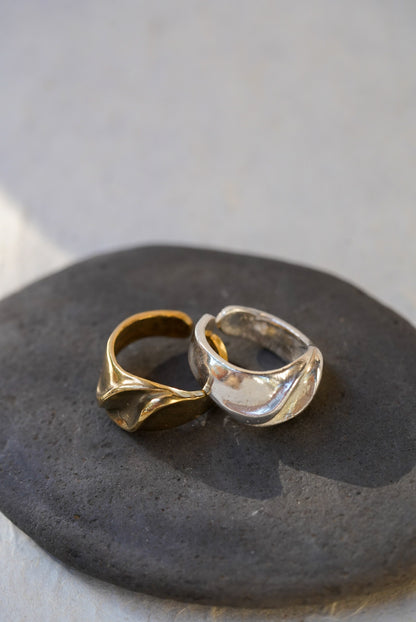 Two textured abstract wave-shaped rings, one bronze and one silver, displayed on a dark slate stone against a light gray background.