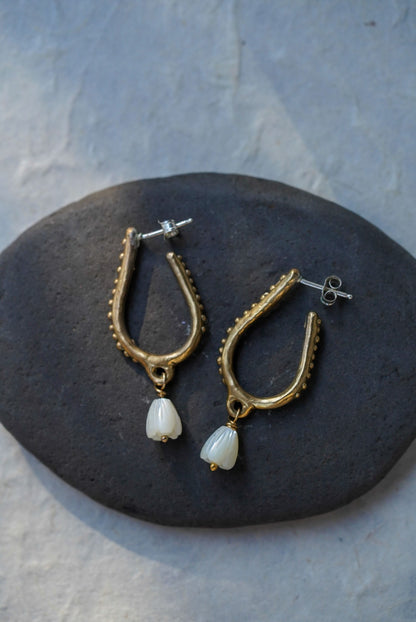 A pair of bronze hoop earrings with hand carved flower beads, displayed on a dark stone against a light gray textured background.