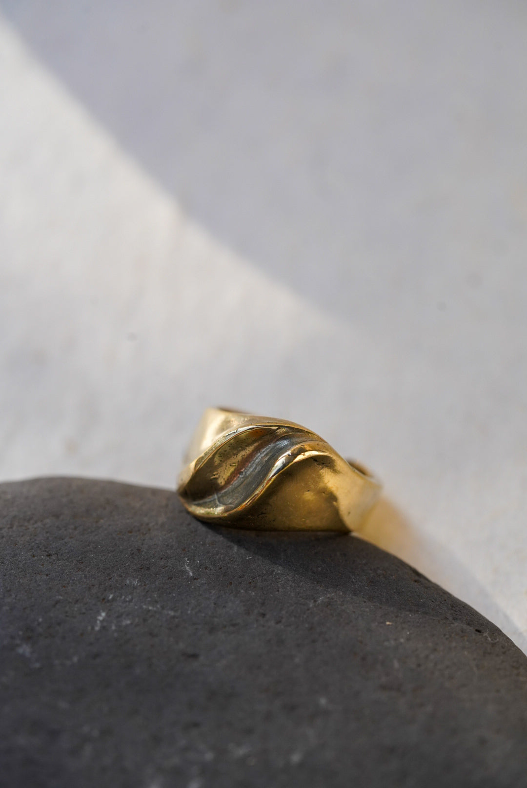 A bronze ring with a twisted, organic wave-like design, resting on a dark textured stone against a light gray background.