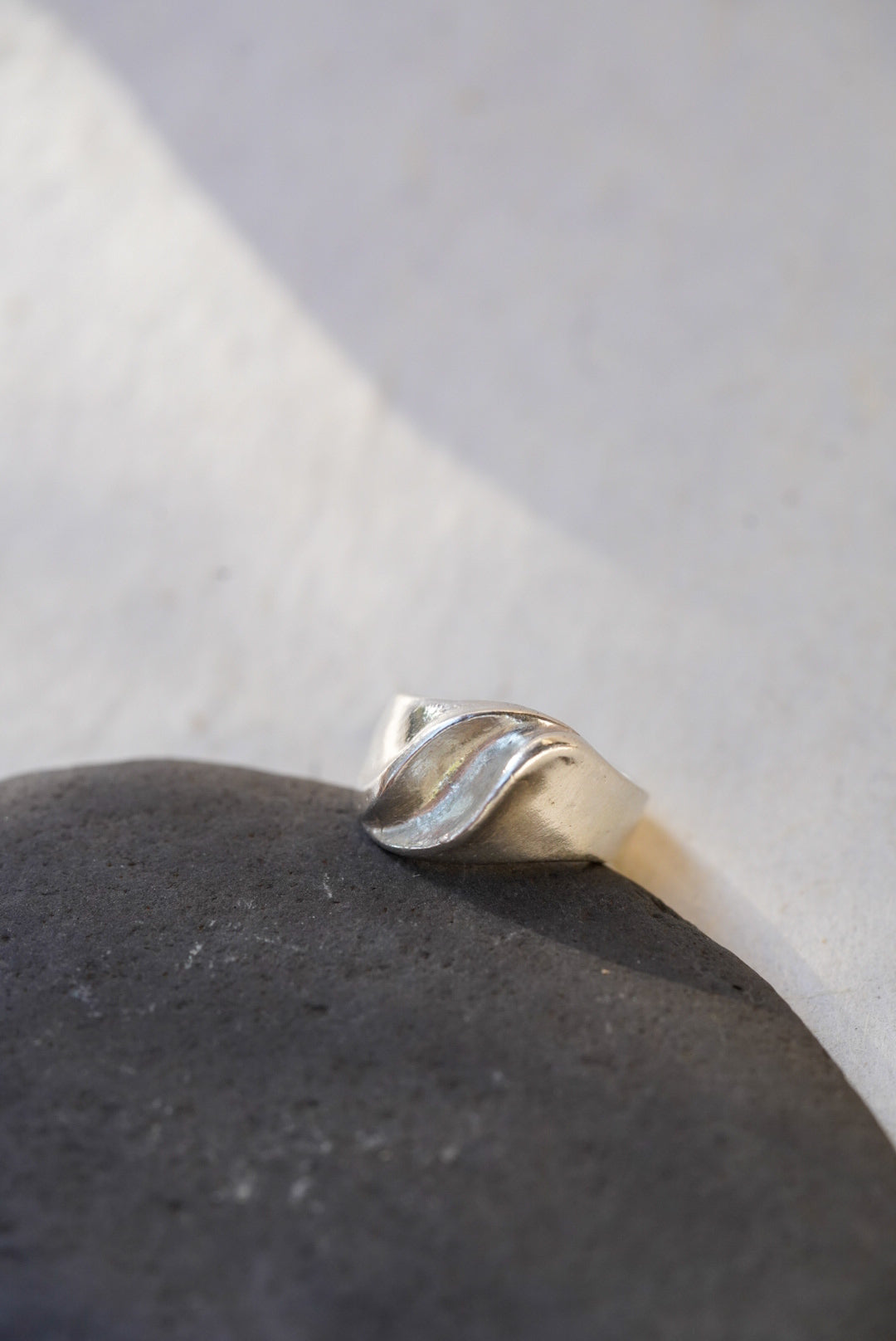 A silver ring with a sculptural, organic wavy band, resting on a dark textured stone against a light gray background.