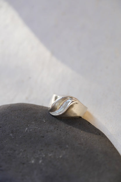 A silver ring with a sculptural, organic wavy band, resting on a dark textured stone against a light gray background.