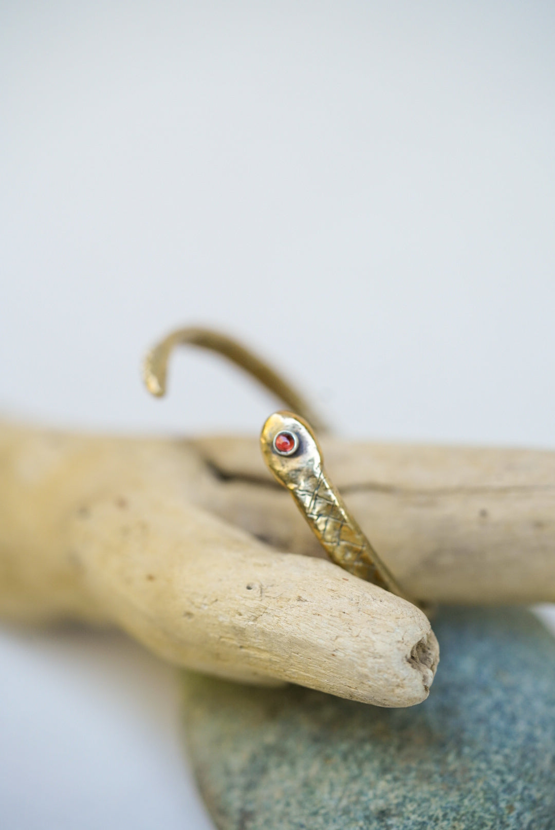 Gold snake-shaped cuff with a red stone on a piece of driftwood against a white background