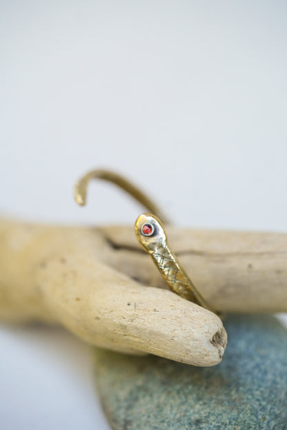 Gold snake-shaped cuff with a red stone on a piece of driftwood against a white background
