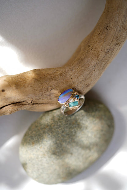 Gold ring with opal and blue stones on a stone surface with driftwood