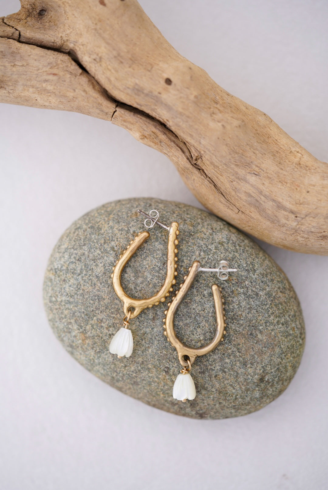 Gold earrings with white pearls on a stone surface with driftwood in the background