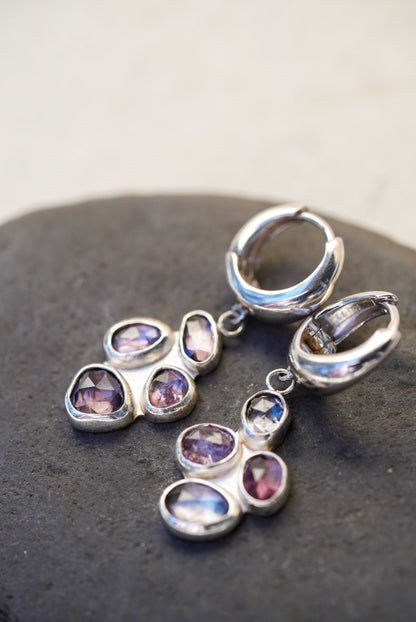 A pair of handmade silver hoop earrings with a cluster of faceted stones in purples and grey, displayed on a dark gray stone against a light beige background.
