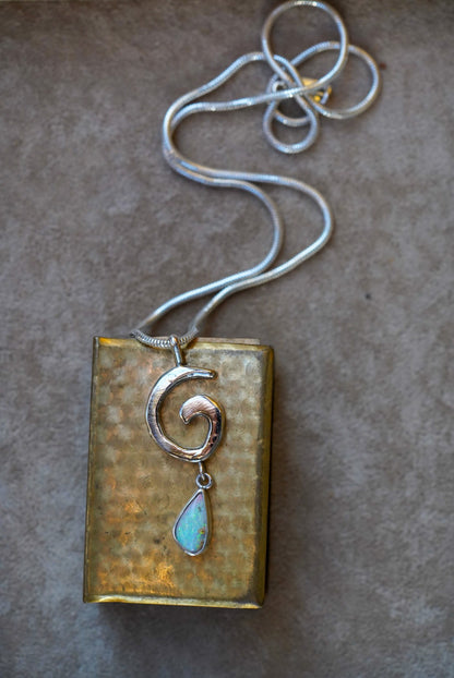 A silver necklace with a spiral pendant and a teardrop-shaped iridescent opal drop, displayed on a dark stone against a vintage gold matchbox on a textured off-white background.
