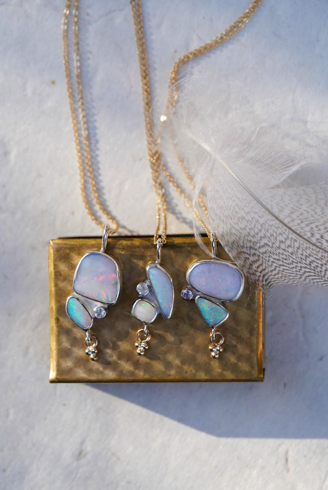 Opal gemstone necklaces in silver settings, displayed on a vintage gold matchbox with a soft white feather background.