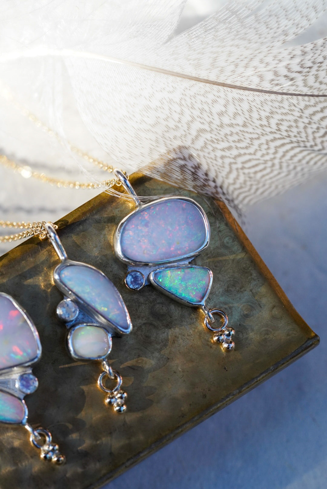 Opal gemstone necklaces in silver settings, displayed on a vintage gold matchbox with a soft white feather background.