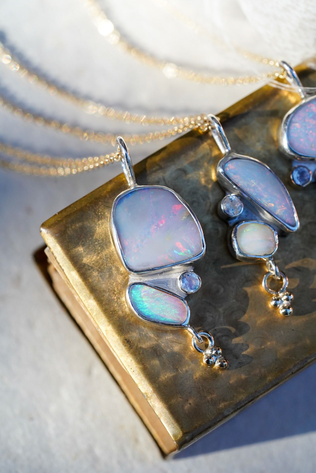 Three Silver necklaces featuring opals and a moonstone along with a small gold charm below, displayed on a textured vintage gold matchbox against a light neutral background.