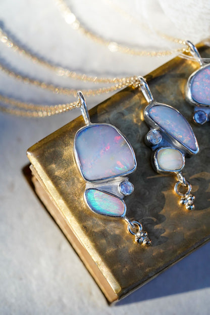 Three Silver necklaces featuring opals and a moonstone along with a small gold charm below, displayed on a textured vintage gold matchbox against a light neutral background.