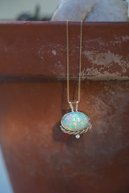 A gold necklace with an oval iridescent opal pendant with a twisted gold accent and small diamond below, hanging on a terracotta background
