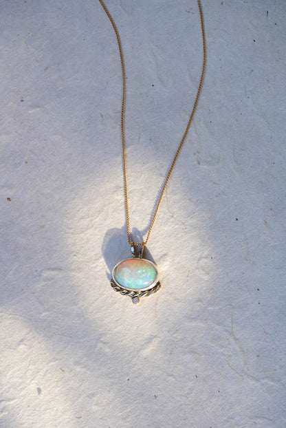A gold necklace with an oval iridescent opal pendant with a twisted gold accent and small diamond below,  against a textured off-white background.