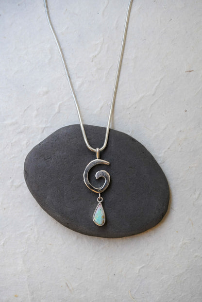 A silver necklace with a spiral pendant and a teardrop-shaped iridescent opal drop, displayed on a dark stone against a textured off-white background.