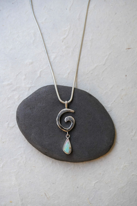 A silver necklace with a spiral pendant and a teardrop-shaped iridescent opal drop, displayed on a dark stone against a textured off-white background.