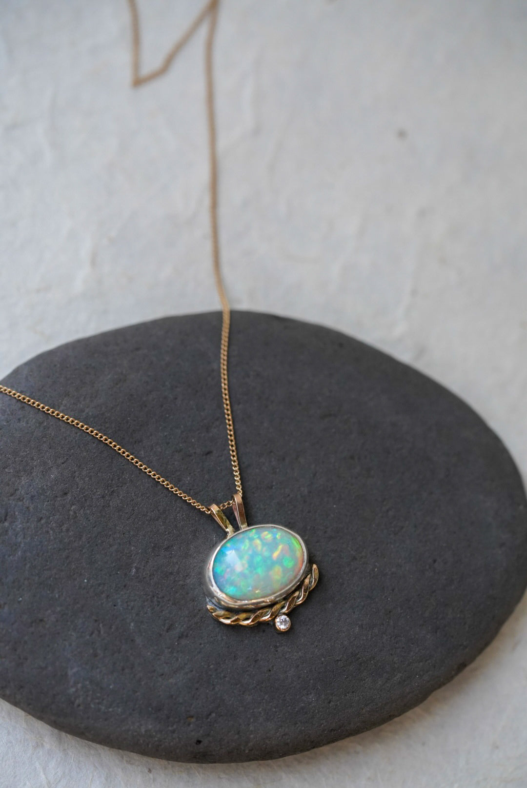 A gold necklace with an oval iridescent opal pendant with a twisted gold accent and small diamond below, resting on a dark gray stone against a textured off-white background.