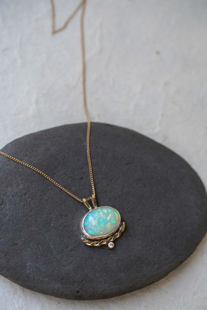 A gold necklace with an oval iridescent opal pendant with a twisted gold accent and small diamond below, resting on a dark gray stone against a textured off-white background.