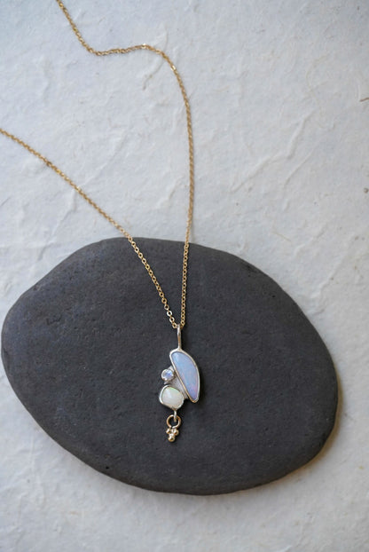 A delicate gold chain necklace with a pendant featuring a curved iridescent opal stone, another small opal stone, and a moonstone, and a tiny dangling gold charm, displayed on a dark gray stone against a light textured background.