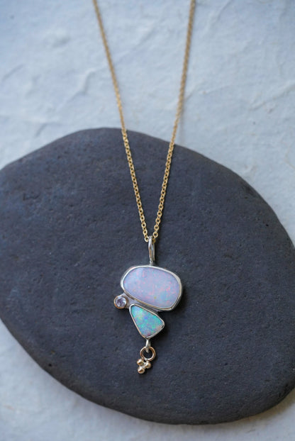 A gold chain necklace with a unique pendant featuring a large opal stone and a smaller light blue-green opal stone alongside a moonstone and a small charm underneath. Displayed on a dark gray stone against a textured off-white background.