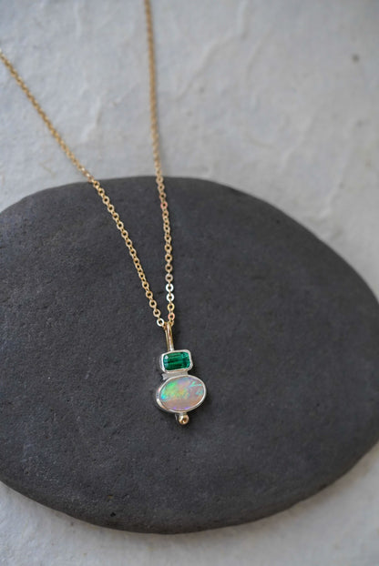 A delicate gold chain necklace with a silver pendant featuring a green rectangular tourmaline and an oval iridescent opal, displayed on a dark gray stone against a light gray background.