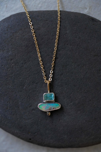 A delicate gold chain necklace with a pendant made of stacked natural stones, one tourmaline and one opal, displayed on a dark gray stone slab against a textured off-white background.