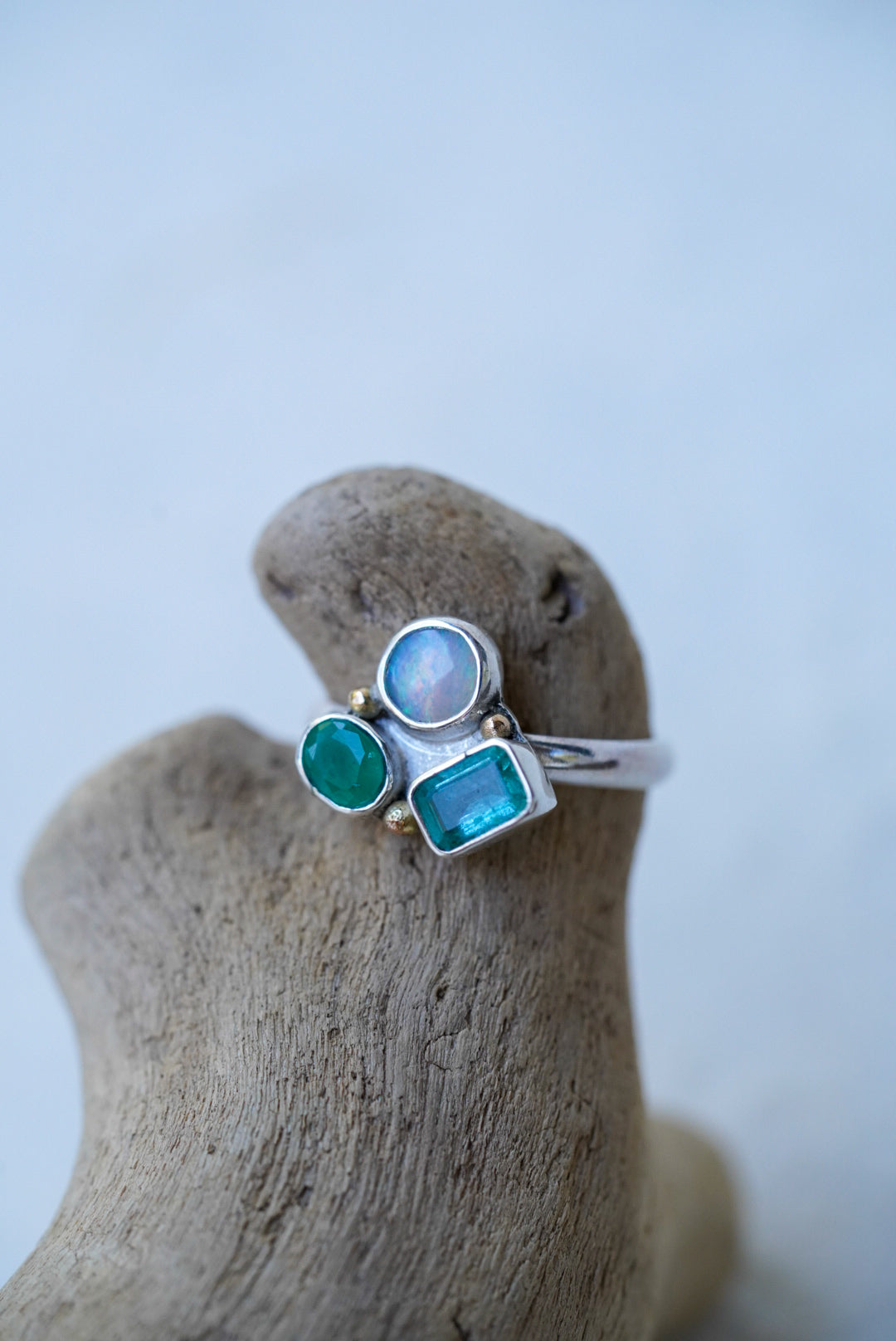 A silver ring with three gemstones (a round opal stone, a round green stone, and a rectangular blue-green stone) displayed on a piece of driftwood against a light background.