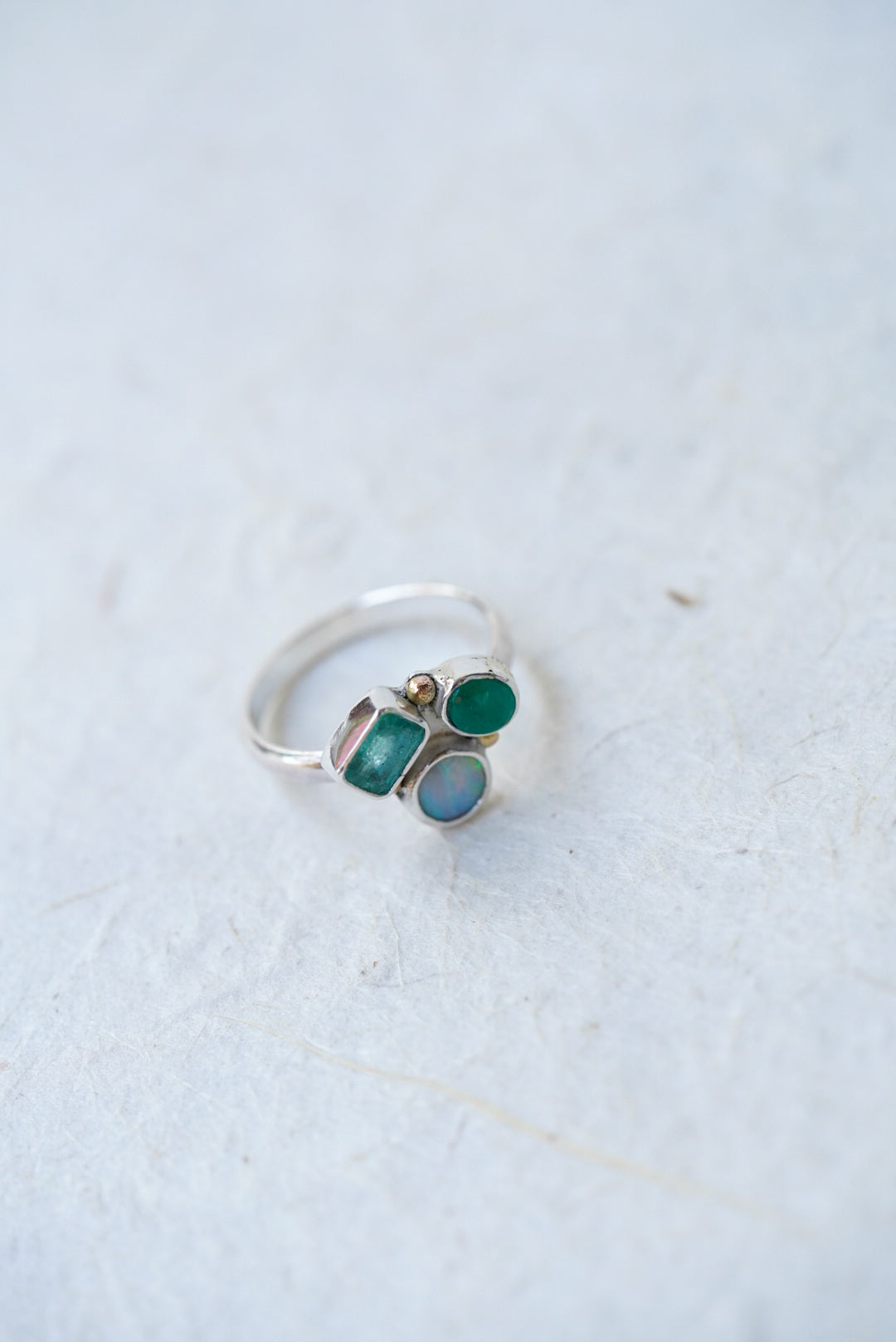 A silver ring featuring three small gemstones in green and light blue, photographed on a light textured surface.