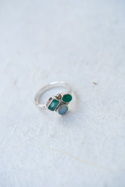 A silver ring featuring three small gemstones in green and light blue, photographed on a light textured surface.