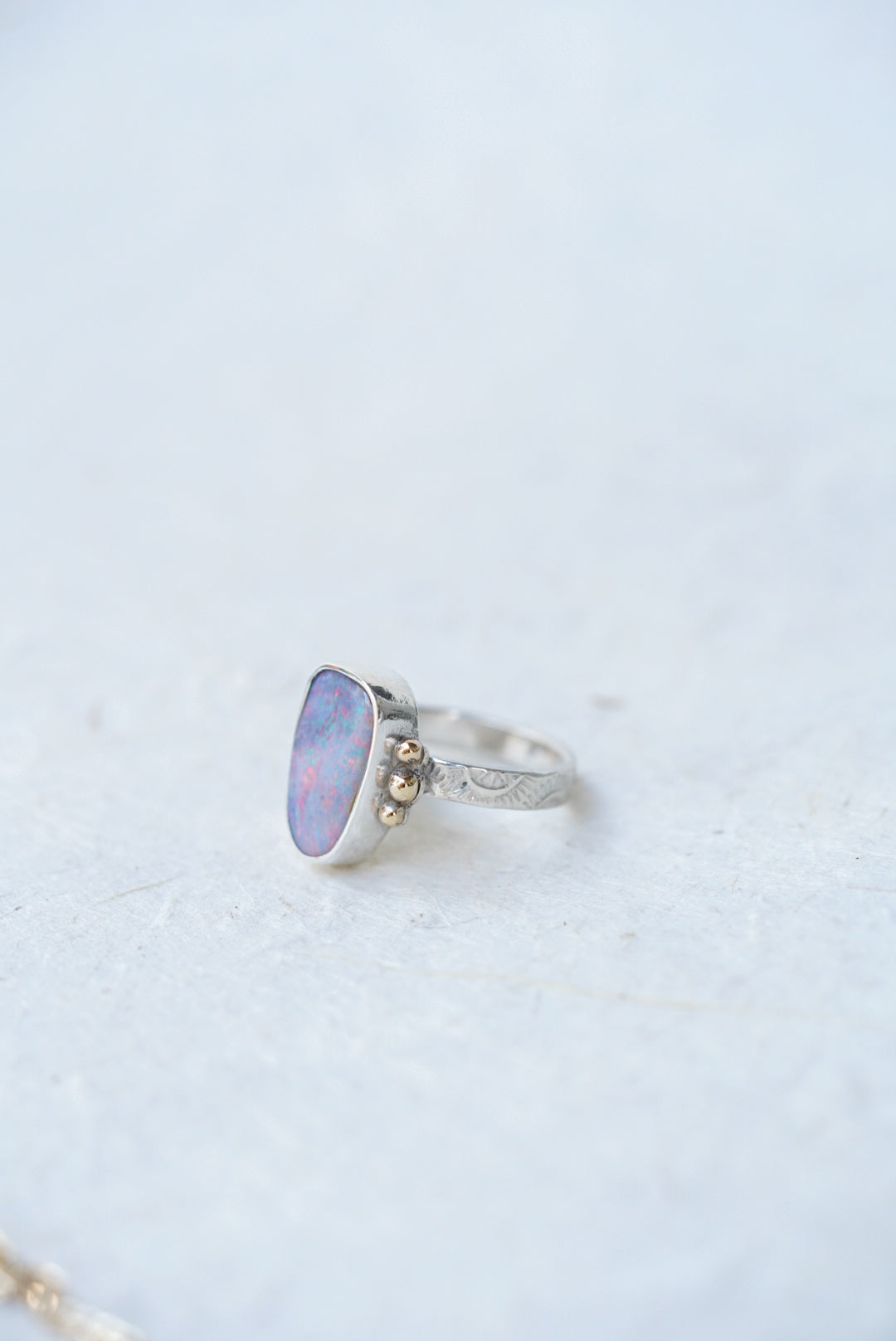 A silver ring featuring a cabochon opal gemstone with iridescent blue, purple, and pink hues, accented by three small gold accents on the band, set against a light textured background.