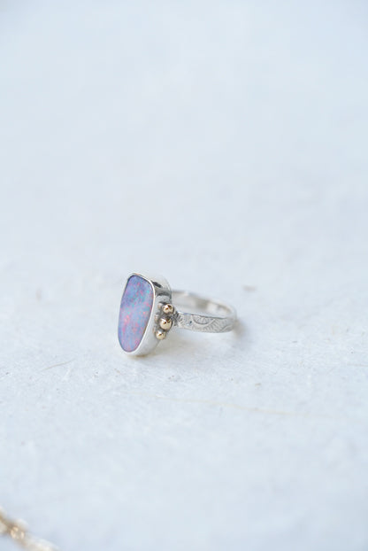 A silver ring featuring a cabochon opal gemstone with iridescent blue, purple, and pink hues, accented by three small gold accents on the band, set against a light textured background.