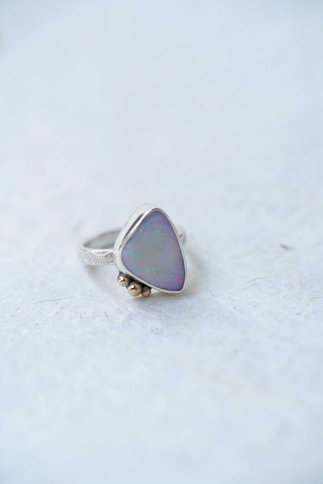 A silver ring featuring a triangular iridescent opal stone with a soft purple and blue sheen, accented by three small gold accents on the stone, set against a light gray textured background.