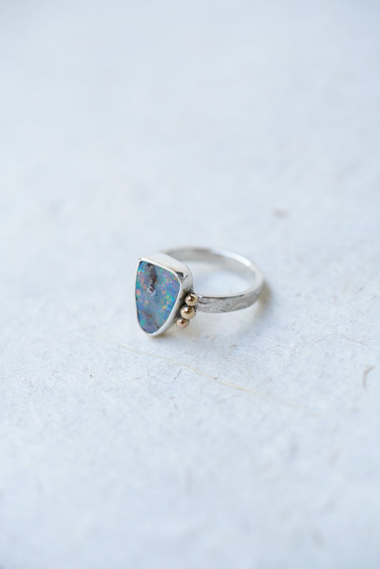 A silver ring featuring a triangular opal stone in a bezel setting, accented with small gold accents on the stone, set against a light textured background.