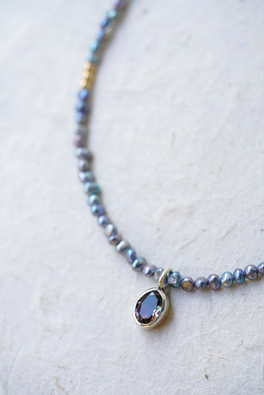 A closeup of a delicate beaded necklace with small iridescent blue-gray pearls, gold spacer accents, and a tourmaline at the center, laid on a light textured surface.
