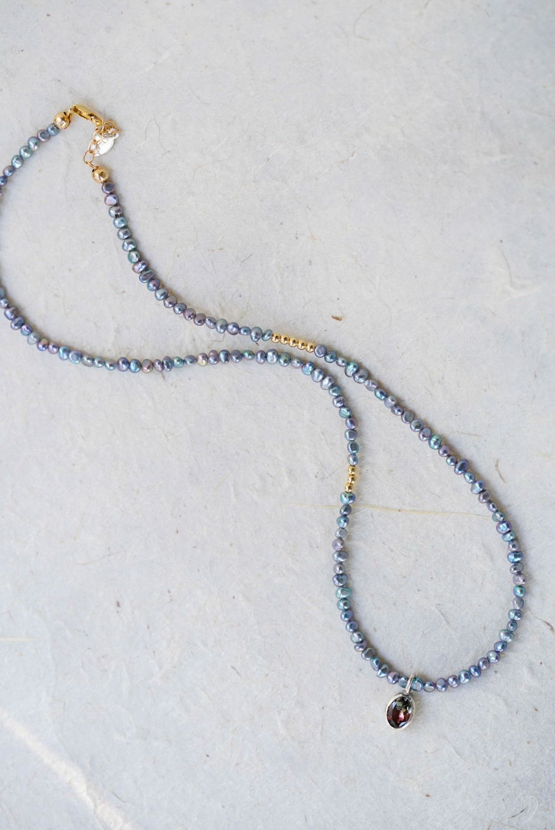 A delicate beaded necklace with small iridescent blue-gray pearls, gold spacer accents, and a tourmaline at the center, laid on a light textured surface.