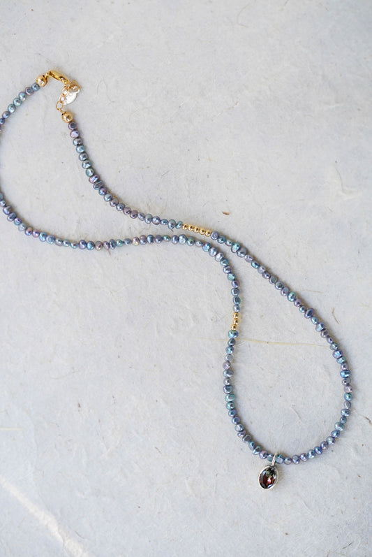 A delicate beaded necklace with small iridescent blue-gray pearls, gold spacer accents, and a tourmaline at the center, laid on a light textured surface.