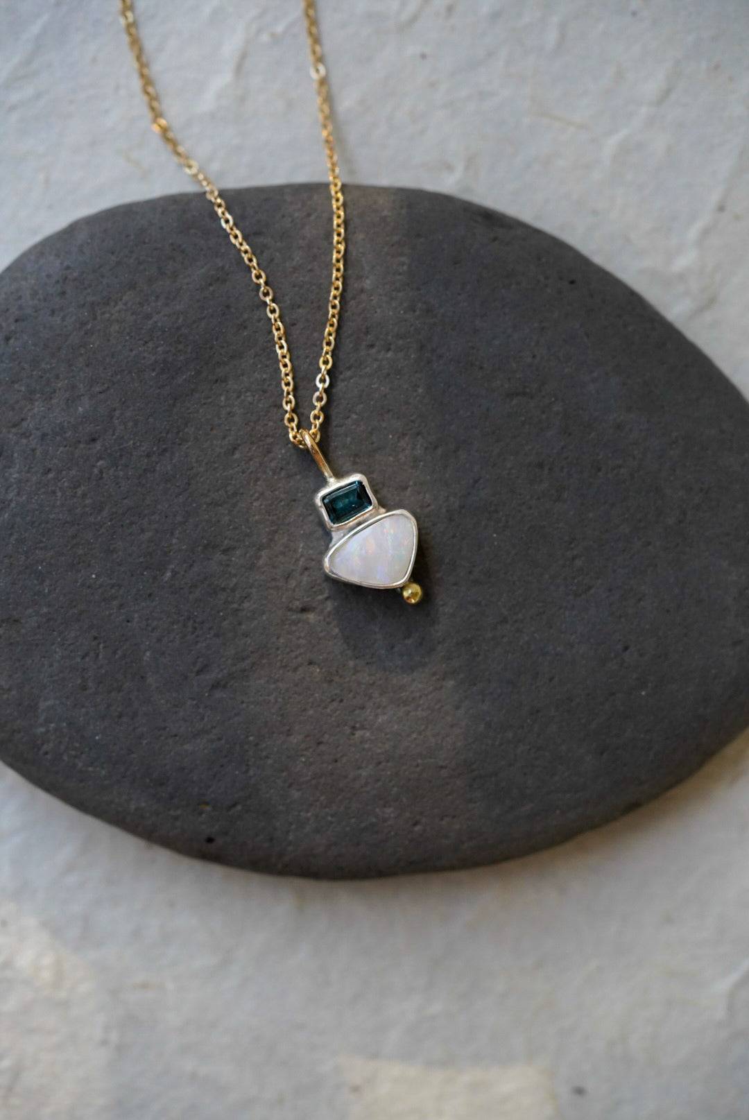 A delicate gold chain necklace with a small silver pendant featuring a white teardrop-shaped opal stone, a tiny dark tourmaline square, and a small gold bead, displayed on a dark gray stone against a light gray background.