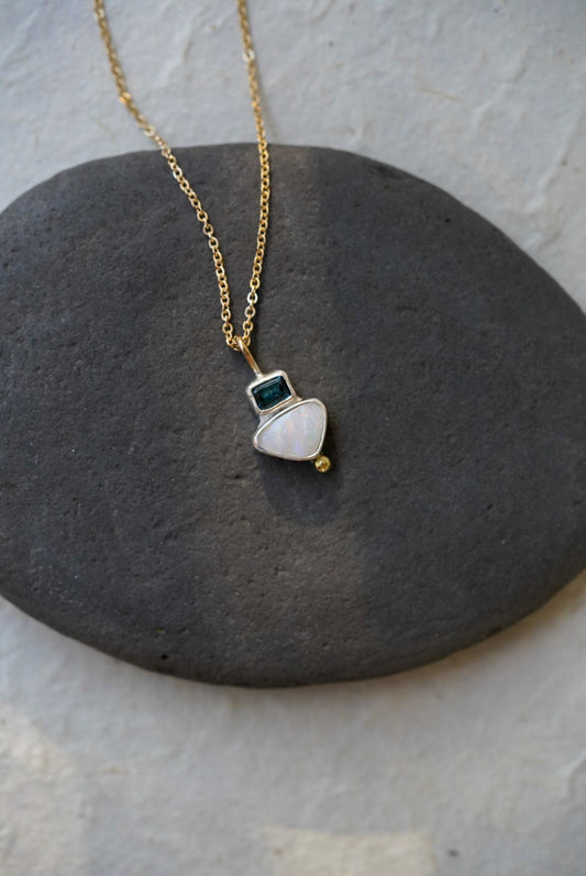 A delicate gold chain necklace with a small silver pendant featuring a white teardrop-shaped opal stone, a tiny dark tourmaline square, and a small gold bead, displayed on a dark gray stone against a light gray background.