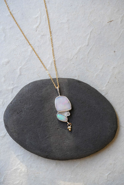 A gold chain necklace with a silver pendant featuring a large iridescent opal and a smaller green-blue opal opal, accented by a moonstone and a dangling gold bead, displayed on a dark gray stone against a light textured background.