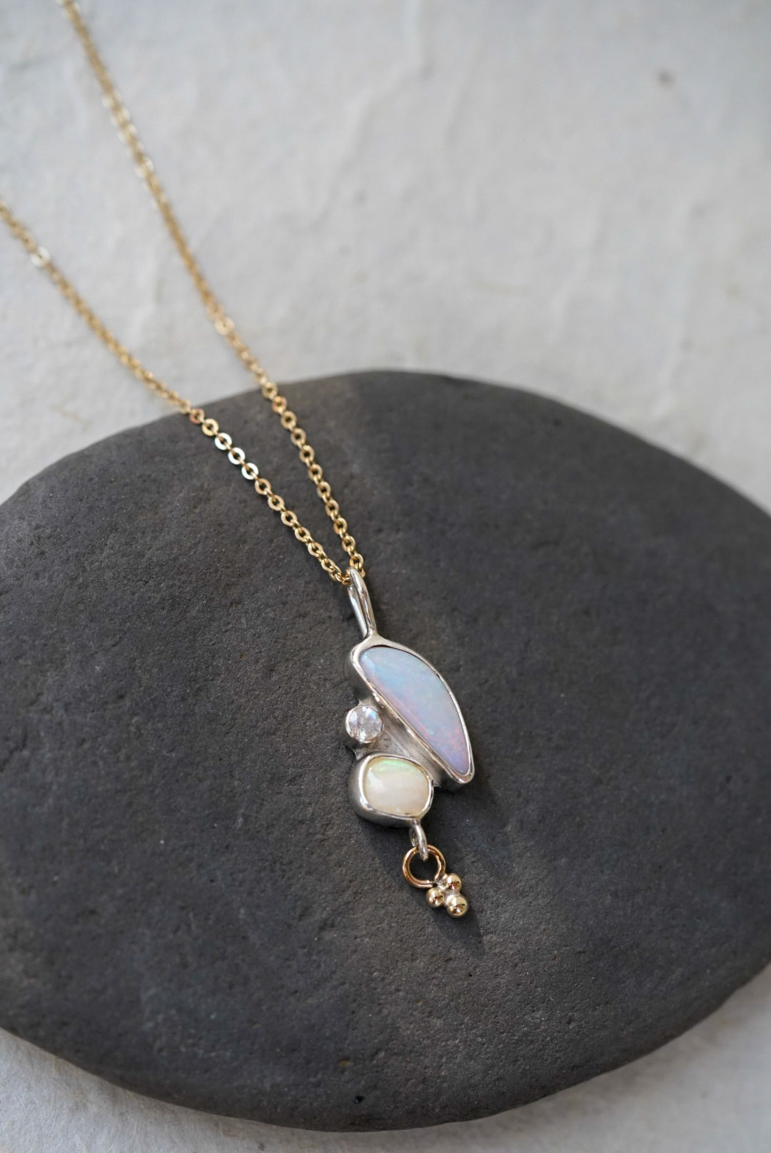 A delicate gold chain necklace with a pendant featuring a curved iridescent opal stone, another small opal stone, and a moonstone, and a tiny dangling gold charm, displayed on a dark gray stone against a light textured background.
