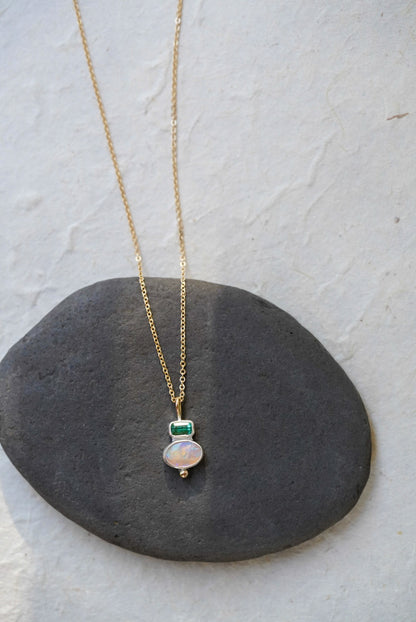 A delicate gold chain necklace with a silver pendant featuring a green rectangular tourmaline and an oval iridescent opal, displayed on a dark gray stone against a light gray background.
